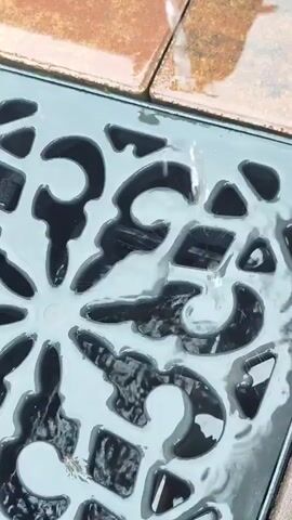 Water droplets are falling onto a decorative metal grate. The grate has an ornate, possibly floral or scrollwork pattern with many openings. The water is falling from the upper right corner of the frame and passing through the grate. The grate is wet, and some of the openings are filled with water. The surface surrounding the grate appears to be made of reddish-brown tiles. The overall scene suggests an outdoor setting, possibly a patio or walkway, where water is draining away.