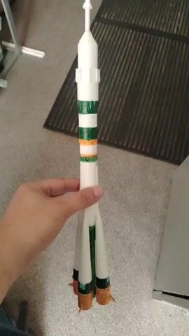 A close-up, handheld shot shows a person holding a white model rocket. The rocket has several green and orange stripes circling its body. It has four boosters at the base, colored green with orange tips. The rocket is held in front of a black mat and a gray carpeted floor. In the background, a wooden cabinet and a metal rack are visible. The lighting suggests an indoor setting, possibly daytime, given the natural light in the background. No text is visible in the frame. The person's hand is the only indication of a human subject. The focus is entirely on the rocket model.