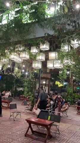 Here is a description of the video:
The video shows an outdoor cafe at night.  The cafe is filled with tables and chairs, and people are sitting at some of the tables. There are many square lanterns hanging from the trees above the tables, providing light. Several employees, dressed in grey shirts and black pants, are cleaning up around the tables. One employee is sweeping, while another is moving a small wooden table. A bicycle is parked on the sidewalk. The sidewalk is paved with red bricks and is next to a busy street.