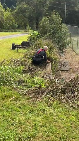 A bald man in a black shirt and black pants with a red harness kneels beside a concrete structure and a dense patch of grass and weeds. He is using a tool, possibly a saw or clippers, to cut something within the greenery. The ground around him is covered in debris, including cut grass and branches. In the background, there is a grassy area, a road, trees, a chain-link fence, and a tennis court. There is also a metal object with wheels lying on its side in the grass to the left. The man appears focused on his task.