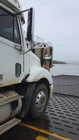 A white semi-truck is parked on the metal grating deck of a ferry in Nizhneleninskoe, Russia. The video begins with a view of the truck's front-left side, showcasing its wheel, fender, and side mirror. The metal deck appears wet, suggesting recent rain or spray. In the background, a wide river flows under an overcast sky, with a large, distant truss bridge spanning its width.

The camera then pans slowly to the right, revealing more of the ferry's structure. White railings, a winch, and chains are visible on the ferry's side. The expansive river continues to be the dominant background element, leading towards the distinct shape of the bridge in the distance. The ferry's deck is uniformly covered with a dark, textured metal grating designed for traction. The scene is set during the daytime under a heavily clouded sky. The context of Nizhneleninskoe indicates this is likely a ferry crossing on the Amur River. No visible text or specific human activity is discernible, but the presence of the truck suggests cargo transport.