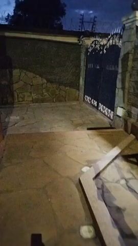 The video shows a partially-lit stone-paved area, possibly a driveway or entrance to a property, at dusk or night. The area is bordered by stone walls and a dark metal gate with decorative elements. A portion of a concrete frame or barrier is visible in the foreground, near the camera. The sky is a dark blue, and some utility wires or electric fencing are visible above the wall in the background. The camera's shadow is visible in the bottom left corner.