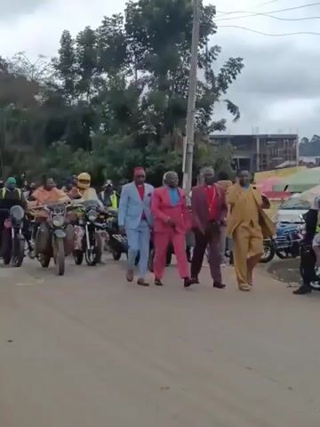The video shows a procession moving along a street. At the front are several men walking in suits of various colors, including blue, pink, and tan. Behind them, a group of people are riding motorcycles and scooters. Some of the riders are wearing helmets and reflective vests. On the side of the road, there are people watching the procession. Trees and buildings are visible in the background, and the sky is overcast. A man in a pink suit is waving at the camera.