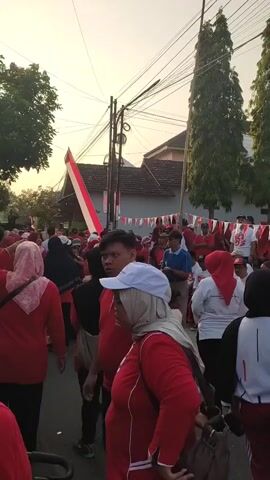 A crowd of people, mostly wearing red and white clothing, are gathered on a street. Some people are holding Indonesian flags, which are red and white. There are trees and buildings in the background. The sky is a pale yellow, suggesting either sunrise or sunset. The overall atmosphere appears to be festive and celebratory. The people are walking and standing, with some interacting with each other. A stage with banners is visible in the distance.