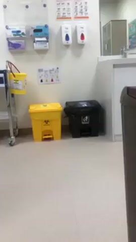 The video shows a clean medical or clinical environment. The floor is light beige with square tiles. On the white wall, there is a clear plastic organizer holding medical supplies like gloves and bandages, and two wall-mounted soap dispensers, one with blue soap and one with red soap. There are also instructional posters on the wall and a metal cart to the left with a small yellow bucket attached. On the floor, there are two trash cans: a yellow biohazard can with a foot pedal and a black trash can with a picture of a recycling symbol. In the background, there is a stainless steel sink and cabinet. A brown object is visible on the right side of the frame, partially obstructing the view. The overall impression is of a sterile and organized setting.