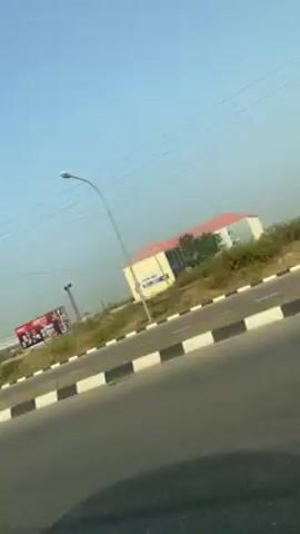 The video shows a roadside view taken from a moving vehicle. The road has a black and white curb. On the side of the road, there are power lines and utility poles. Further in the background, there are buildings and a billboard. The sky is light blue.
