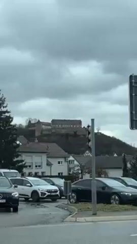 The video shows a view from inside a vehicle at an intersection on a cloudy day. In the distance on a hill sits a castle or fortress. There are multiple cars visible in the foreground, waiting at a traffic light. The camera is static, capturing the scene through the vehicle's window. The environment appears to be a town or residential area with houses interspersed with trees. The overall tone is neutral and documentary-like, simply recording the scene as it unfolds.