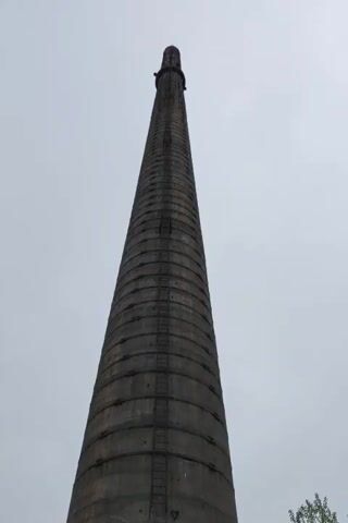 The video shows a tall, cylindrical concrete smokestack from a low angle, looking upwards. The smokestack is weathered and has a ladder running up its side. At the base of the smokestack, there is an opening with the word "ОПАСНО" (DANGER) spray-painted above it. The surrounding environment appears to be an abandoned industrial area, with overgrown vegetation, debris, and puddles of water on the ground. The sky is overcast.