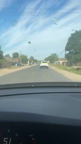 A view from inside a vehicle, looking forward through the windshield. The road ahead is paved and has white lane markings. Several cars are visible on the road, including a white police car with flashing lights and the word "POLITIA" on the back, in front of the camera. There are trees and buildings on either side of the road, and a blue sky with some wispy clouds is overhead. The camera appears to be moving, as indicated by the slight blur in the image. There are several dark spots visible on the windshield, which may be debris or damage.