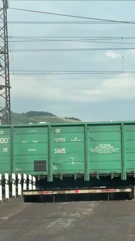A train is traveling across a railway crossing. The train cars are green and blue and are covered in text and numbers. In the background, there are power lines and rolling green hills under a cloudy sky.