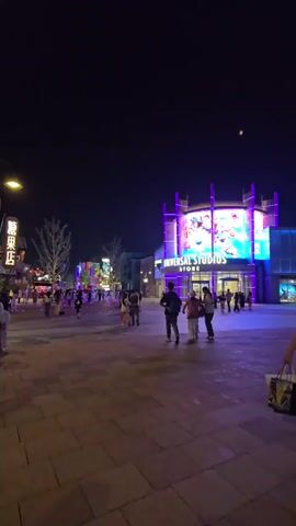 The video shows a nighttime view of a plaza area with several buildings and a crowd of people. On the right side of the frame, a large building has "UNIVERSAL STUDIOS STORE" displayed prominently on its facade, illuminated with purple and blue lights. A large screen above the store entrance displays a colorful image.

To the left, there are several other buildings with illuminated signs, including one that appears to say "Candy Smith". The plaza is paved with square stone tiles. Numerous people are walking around the plaza, some in groups, some individually. The overall lighting is provided by streetlights, building lights, and the illuminated signs and screens, creating a vibrant, festive atmosphere. The sky is dark, indicating it is nighttime, and a faint moon or celestial object is visible.