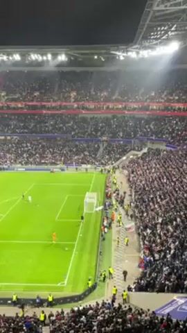 The video shows a soccer match in a large stadium at night. The stadium is filled with spectators. The field is green and the players are in various locations on the field. The crowd is dense, with people filling the seats and walkways. Bright lights illuminate the field. People are cheering.