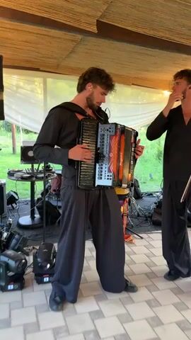 A man with a beard and curly hair plays an accordion. He is wearing a black shirt with wide sleeves and black pants that are baggy and flare out at the bottom. He is on a stage, and there are lights and other musical equipment around him. In the background, there is a ceiling made of bamboo, and some white curtains. There is also some greenery visible outside. Another person is partially visible on the right side of the frame. The man is looking down at the accordion as he plays.