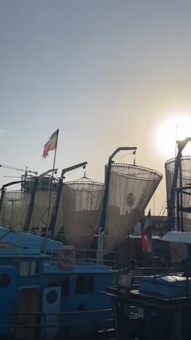 The video shows a close-up of a man on a boat, talking on his phone. He is wearing green shorts and is bare-chested. He is leaning against the boat's cabin. In the background, there are several other boats and fishing nets. The sun is setting, casting a warm glow over the scene.