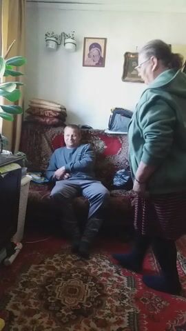 A man is sitting on a couch in a living room with a patterned rug. An older woman is standing next to the couch. There are framed pictures on the wall, and a houseplant is in the corner of the room.