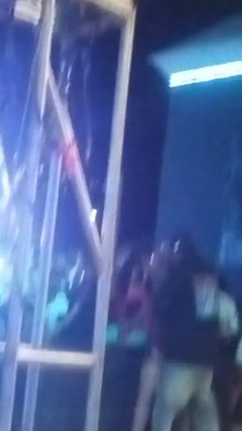 The video appears to be shot from a lower angle, possibly from behind scaffolding at an event. Initially, bright stage lights and silhouetted figures are visible. The focus then shifts to a group of people standing near a wall with writing on it. The figures are blurred, but it's possible to make out articles of clothing, such as a black jacket, blue jeans, a white top, and a striped shirt. It is difficult to identify specific actions or interactions in the video.