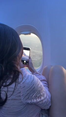 Here is a concise description of the video:

The video shows a woman on an airplane taking a picture with her smartphone of the view out the window. The airplane wing is visible through the window along with the city below at sunset. The light is bright and warm.