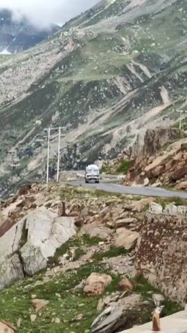 A truck drives along a winding road in a mountainous landscape. The road is paved and appears to be two lanes wide. The truck is white with colorful decorations on the front. The surrounding environment is characterized by steep, rocky slopes covered in patches of green grass and sparse vegetation. In the distance, towering mountains with some snow or clouds are visible. The sky is partly cloudy, with sunlight intermittently breaking through. Power lines and poles are also present alongside the road. The overall scene depicts a remote, high-altitude road traversing a rugged and scenic mountainous terrain.