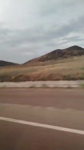 A first-person perspective video shows a landscape with hills under a cloudy sky. The foreground is blurred, indicating motion, and a white line from a road is visible. The hills in the background are covered in dry grass and sparse vegetation. The overall impression is of a rural or undeveloped area, possibly experienced from a moving vehicle.