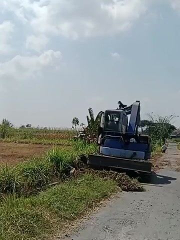 An excavator is being operated on the side of a paved road next to a field of tall green plants. The excavator is blue and white and is digging into the ground, moving dirt. The bucket of the excavator is actively scooping and dumping soil. The field appears to be a rural agricultural area with a few small structures visible in the background. The sky is cloudy with patches of blue.