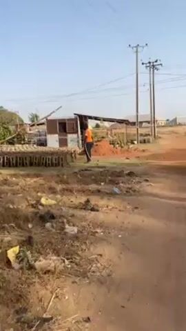 A person in an orange shirt and dark pants is standing on a dirt and grassy field. The person is facing away from the camera and holding a long, thin, light-colored object, possibly a hose or a rope. To the person's left is a stack of hollow rectangular blocks. Behind the person is a pile of red dirt and a dilapidated structure with a corrugated metal roof. The environment includes dry grass and scattered trash on the ground. In the background are utility poles and indistinct buildings. The sky is light blue. The video appears to be shot on a sunny day.