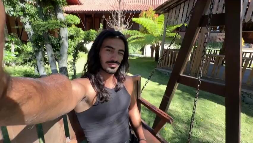 The video shows a man with long, dark hair sitting on a wooden swing in an outdoor setting. He is wearing a black tank top. He is holding up his right arm, likely to take a selfie. The background shows a lush green lawn, trees, and wooden buildings. The lighting suggests it is daytime and sunny. The man is looking towards the camera with his eyes closed initially, then opens them and smiles. He seems relaxed and enjoying the environment.