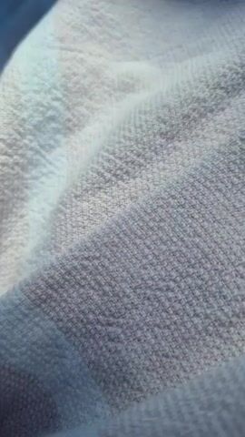 A close-up shot focuses on a white, textured fabric, possibly a towel or blanket, with thin vertical stripes. A tiny black insect, likely an ant, is visible on the fabric. In the background, a green clothespin is attached to the fabric, and out-of-focus green foliage and brown ground are present, suggesting an outdoor environment. The lighting indicates it is daytime. The camera remains mostly stationary, with slight movements and focus shifts.