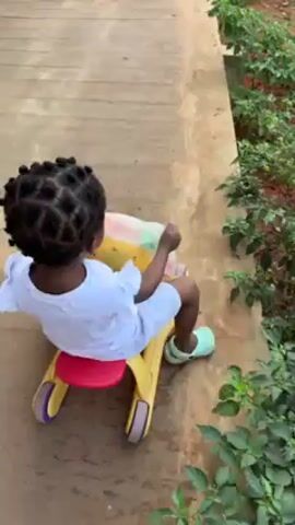 A toddler in a white dress and green sandals sits on a yellow and red ride-on toy. The toddler is facing away from the camera, on a concrete walkway or path, with some greenery visible to the right. The child begins to move forward, rolling on the toy. The camera follows behind. A blue bicycle is visible to the left as the child moves away from the camera.