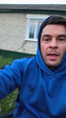 This is a selfie-style video taken outdoors in what appears to be a rural setting, possibly a backyard or dacha in Yaganovka, Russia. The primary subject is a man with short brown hair and a light complexion, wearing a bright blue hooded sweatshirt. He is smiling and looking directly at the camera, appearing to be in a good mood.

The background features a light-colored, horizontally-sided building with a white-trimmed window. Green grass and foliage are abundant, indicating it is likely daytime, possibly late afternoon or early evening given the lighting. To the right of the man, a metal barbecue grill with a fire burning inside is visible, suggesting the activity taking place is cooking or preparing for a barbecue. Sparks can be seen emanating from the fire in some frames. A white fence and darker soil, possibly a garden plot, are also visible in the background.

The overall atmosphere is relaxed and cheerful, with the man's smile and the presence of the barbecue contributing to a sense of leisure and enjoyment. No visible text is present in the video.