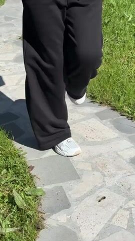 A person wearing white shoes and black pants walks on a stone pathway with grass on either side. The camera follows their feet as they walk. The person also holds a phone in their left hand.
