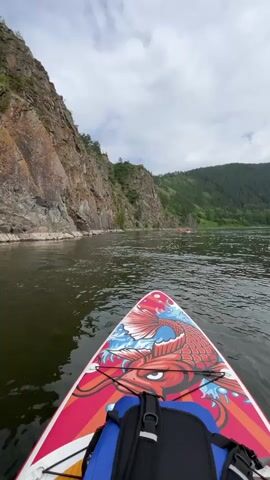 A first-person perspective shot shows the front of a paddleboard with a colorful koi fish design. A blue backpack is secured to the board. The paddleboard is on a body of water, likely a river, with a rocky cliff face on the left and a dense, green forest covering rolling hills in the distance on the right. The sky is overcast with scattered clouds. Several small red objects, possibly other paddleboards or kayaks, are visible further down the river in the distance. The water is calm with some ripples. The overall scene depicts a serene natural environment, possibly during an outdoor recreational activity like paddleboarding or kayaking.