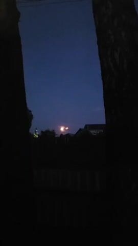 The video shows a dark night scene with a bright, out-of-focus moon. The moon appears as a large, luminous orb with two smaller, blurry circles to its right. Silhouettes of trees frame the left and right sides of the image. In the background, dark shapes suggest houses or buildings. The sky is a deep, dusky blue, fading to black at the top. A blurry, horizontal line near the bottom may indicate a fence or railing. The overall impression is of a nighttime view through trees, with the moon as the focal point.