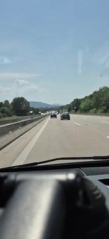 This media file captures a view from inside a vehicle driving on a highway, likely in Germany given the location context of Malsch. The scene is set on a sunny day with clear blue skies and some scattered clouds, suggesting fair weather. The highway appears to have at least two lanes in the direction of travel, with a concrete barrier separating it from the opposing lanes.

The primary subject is the highway traffic. Several cars and trucks are visible, driving in the same direction. The closest car ahead is a dark-colored hatchback, followed by other vehicles at varying distances. Further down the road, a significant traffic jam is forming or in progress, with numerous vehicles slowed or stopped. The presence of trucks indicates this is a major transportation route.

The setting includes lush green trees and vegetation lining the sides of the highway, with a gentle incline of a hill in the distance. The time of day is likely midday, given the bright sunlight casting shadows. The overall mood appears to be one of typical highway travel, potentially with a hint of frustration due to the developing traffic congestion. No visible text is present in the media file. The perspective suggests the media was captured by someone driving or being driven, with the dashboard of the vehicle slightly blurred in the foreground.