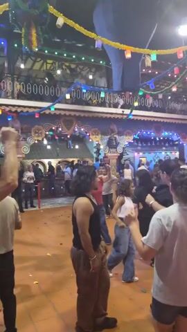 The video shows an indoor gathering with many people, possibly a party or festival. The environment is decorated with colorful lights and streamers hanging from the ceiling. People are seen dancing and socializing in the main open area. In the background, there are elevated platforms and structures with more people and decorations. Confetti is scattered on the floor, suggesting a celebratory atmosphere. The lighting is dim but illuminated by the decorative lights.