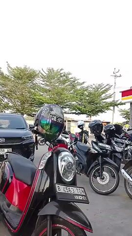 The video displays a row of motorcycles parked outside what appears to be a convenience store. The foremost motorcycle is red and black, with a black helmet placed on its handlebars. This helmet has a reflective, iridescent visor and displays some text that is illegible. Behind this motorcycle are several other black motorcycles, all with helmets resting on them. In the background, a black SUV is visible, and beyond that, trees and the awning of the convenience store, which has red and yellow accents. The sky is overcast and white. The overall impression is a brief, static shot of a parked motorcycle convoy or gathering.