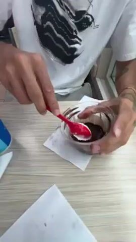 A person with a tattoo on their right forearm is sitting at a light brown table, stirring chocolate pudding in a clear plastic cup with a red plastic spoon. The cup is sitting on a white napkin. A second white napkin is also visible on the table. The person is wearing a white t-shirt with a black and white graphic design on it. A blue and white cup is slightly visible on the left side of the frame.