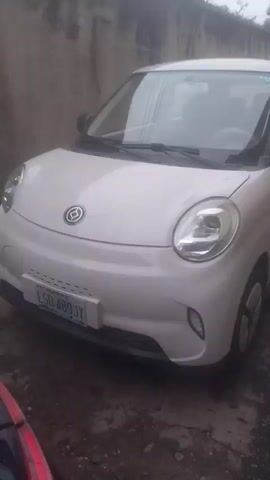 The video shows a light-colored electric car. The camera pans across the exterior and then the interior of the vehicle. The exterior shots focus on the front of the car, including the headlights, grille, and license plate which reads LSD-489JY. The interior shots reveal the steering wheel, dashboard with a screen, and seats. A water bottle is visible on the passenger seat. The environment appears to be an outdoor setting with a wall in the background.
