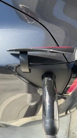 The video depicts a gray Tesla, the charging port lid open, revealing a green indicator light. A charging cable is plugged into the charging port. The car is parked next to a row of charging stations, situated on a concrete curb with a sidewalk.  A person is visible in the car's reflection. The camera moves slightly, providing a view of the car's rear and the charging stations. The video concludes with the person mentioning "30 minutes wait".