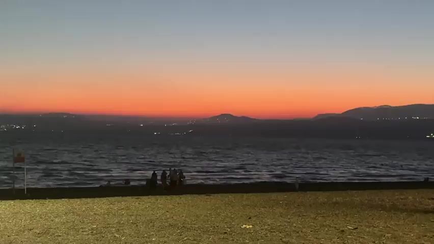 The video shows a beach scene at dusk. The sky is a gradient of blue at the top, fading into orange and then a deeper red near the horizon. In the distance, across a body of water, are hills or mountains, with city lights visible on them. The water is dark and has small waves. On the beach, in the foreground, is a gravelly area. Further back, closer to the water, is a strip of dark sand where a few people are gathered. One person is standing near a pole with a red and white flag. The people are indistinct figures, and their actions are not clear, but they appear to be standing or sitting. The lighting suggests it is late evening or early morning, with the sun having set or not yet risen.