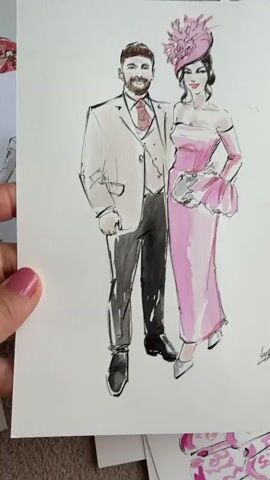 The video shows a hand holding a watercolor painting of a man and a woman in formal attire. The man is wearing a suit, vest, and tie. The woman is wearing a pink dress and a matching hat. The painting is on a white background. There are other pieces of paper and art visible around the painting. The hand has pink nail polish.