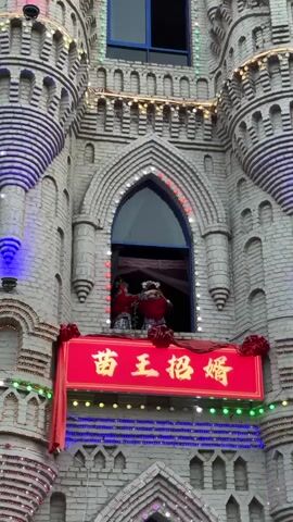 The video shows a festive building that resembles a castle, adorned with colorful lights and what appear to be red ribbons or bows. In an arched window of the castle, two individuals dressed in matching red outfits are performing some kind of dance or ritual. They are facing each other and moving their arms. Below the window, a large red banner with yellow Chinese characters is prominently displayed. The building's facade is made of grey bricks and features decorative architectural elements. The overall impression is of a celebratory event, possibly a wedding or festival.