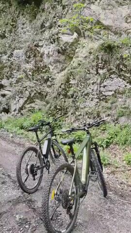 The video captures a scenic view of a winding dirt road surrounded by lush green trees and a rocky hillside. The road is slightly uneven, with some loose gravel visible. On the right side of the road, two bicycles are parked. The hillside on the left is covered with green vegetation, while the right side features a steep, rocky incline with some patches of moss and sparse greenery. A thin stream of water trickles down the rocky hillside on the right, creating a small waterfall effect. The overall scene suggests a tranquil outdoor setting, possibly a nature trail or a mountain path.