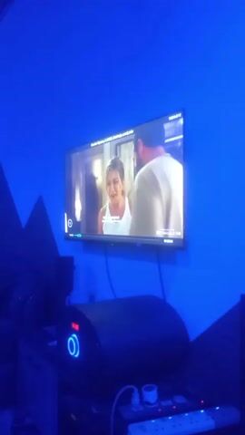 The video shows a television playing in a room with blue walls. On the TV, Jennifer Aniston and a bearded man are visible, and the screen later shifts to a person with sunglasses. Below the television is a black, cylindrical object with a blue ring on it, resting on a table with a power strip. Music plays in the background.
