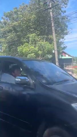 The video opens with a shot of a person riding a red scooter. The scooter is moving along a road with trees and buildings on either side. The sky is blue with some clouds. The camera then pans to the right, showing a white van passing by. Next, the video shows a close-up of a truck with an advertisement for Indomie instant noodles. The advertisement features an image of a bowl of noodles with the text "Soto Koya Pedas Dower." Finally, the video shows a black car parked on the side of the road, with a man sitting on a chair in front of a small shop. The shop appears to be a roadside stall selling various items.