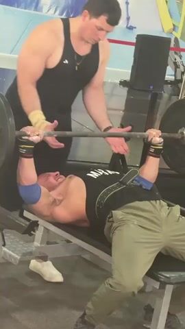 A man is performing a bench press on an incline bench with a spotter standing behind him. The man on the bench is wearing a black tank top, green pants, blue elbow sleeves, and wrist wraps. He is holding a barbell with weights on both ends. The spotter is wearing a black tank top and has his hands on the barbell. The man on the bench begins to struggle with the weight and the spotter intervenes to help him. The man on the bench then pushes himself up off the bench and sits upright, looking distressed. The environment appears to be a gym with other equipment and people in the background.