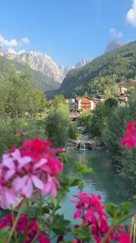 The video captures a scenic view of a village nestled in a valley, surrounded by lush green mountains. In the foreground, vibrant pink flowers are in soft focus, framing the turquoise water of a river flowing through the scene. A small waterfall is visible in the middle ground, cascading down rocks. The village itself consists of traditional houses with orange roofs. The sky is clear and blue with a few scattered clouds. The overall impression is one of natural beauty and tranquility.