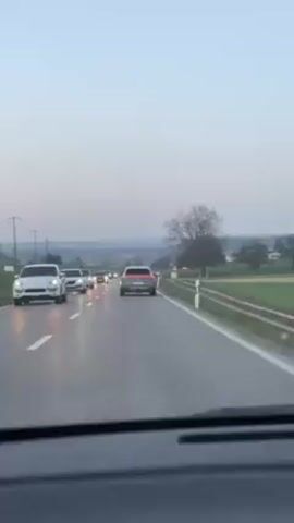 The video shows a car's dashboard view of a roadway populated with several cars. The roadway is grey with white lines denoting the traffic lanes. In the distance, a tree without leaves stands next to a green field. There are other trees and buildings in the distance. Utility poles can be seen in the distance. The sky is a pale, slightly cloudy blue.