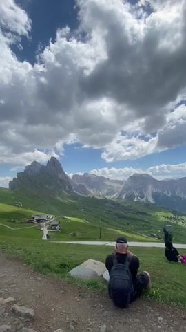 The video shows a panoramic view of a mountainous landscape under a partly cloudy sky. In the foreground, on a dirt path, a person wearing a black hat and a backpack is sitting facing the mountains. To their right, several other people are sitting on the grass, some facing the mountains and one person is doing a handstand. Further in the background, a man in a green and black plaid shirt, black shorts, a hat, and hiking boots is standing and facing away from the camera, looking at the mountains. The landscape features rolling green hills, scattered buildings, and jagged mountain peaks in the distance. The camera pans slightly to the right, showing more of the people sitting on the grass and the expansive mountain range.