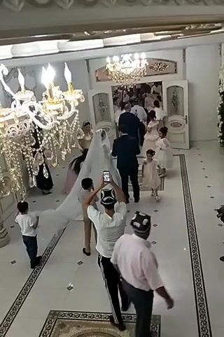 The video captures a wedding ceremony taking place in a large, well-lit indoor hall. The hall features ornate chandeliers and decorative elements, suggesting a formal setting. A bride in a long white wedding dress with a train is the central figure, accompanied by a groom in a dark suit. Several other individuals are present, including guests, children, and possibly wedding party members, some of whom are wearing traditional hats. The group appears to be moving towards an entrance or a designated area within the hall, possibly for a reception or ceremony. The floor is tiled and features decorative patterns. The overall atmosphere seems to be one of celebration.