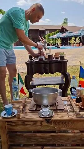A man in a teal t-shirt and grey shorts is standing outdoors and operating a manual espresso machine. The machine is on a dark, ornate table. In front of the table is a wooden display with two tiers. On the top tier, there is a large metal pot, a glass with clear liquid, and a metal object that looks like a miniature samovar, possibly for heating water. On the bottom tier, there is a small blue cup with white polka dots, a few coffee beans, and a spoon. To the left of the wooden display, there is a bottle of water and an Italian flag. To the right of the display, there are more coffee beans, a spoon, another cup, and a Romanian flag. In the background, there is a swimming pool, a building with a red roof, and outdoor seating areas with umbrellas. The man is focused on the espresso machine, and his hands are engaged in operating it. The scene appears to be a demonstration or a specialized coffee-making setup in an outdoor setting.